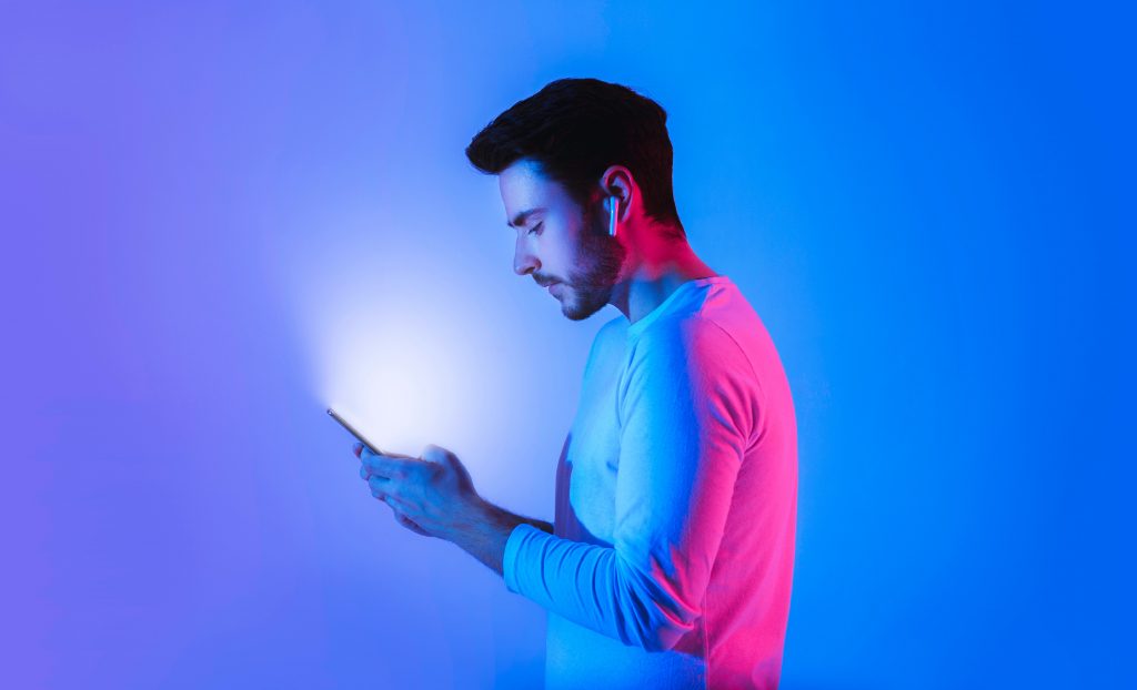 Vue de profil d’un homme jouant sur son smartphone portant des ecouteurs sans fil sous des lumiere neon bleu et rose