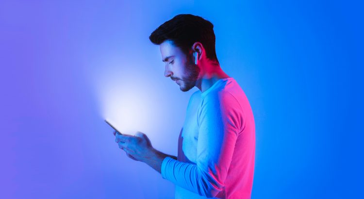 Vue de profil d’un homme jouant sur son smartphone portant des ecouteurs sans fil sous des lumiere neon bleu et rose