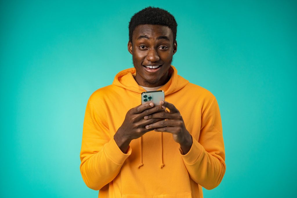 Un jeune homme tenant son smartphone entre les deux mains souriant tout en fixant la camera 