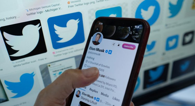 Photo d’un smartphone tenue dans un main affichant le compte Twitter d’Elon Musk. Le fond de la photo montre plusieurs logos de Twitter de différentes couleurs