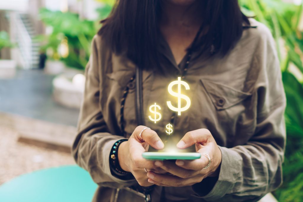 Une femme en train de manipuler son smartphone avec des symbole dollars doré et brillants au-dessus du telephone 