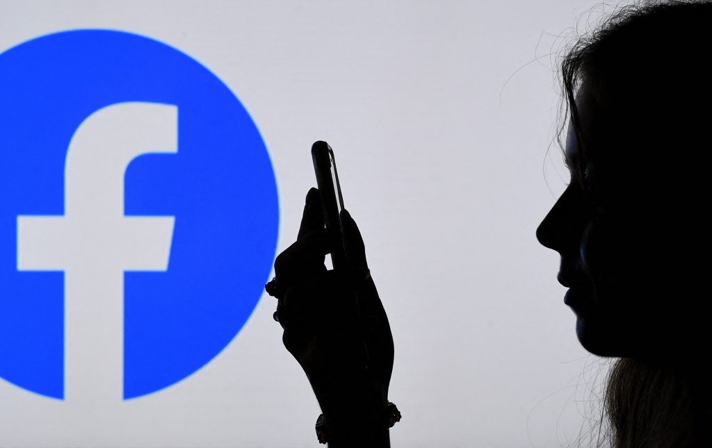 Silhouette d’un profil d’une femme regardant son smartphone devant un grand logo de Facebook 