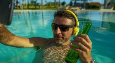 Un homme a lunettes solaires faisant un selfie dans la piscine, un casque aux oreilles et une bouteille de biere dans la main
