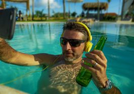 Un homme a lunettes solaires faisant un selfie dans la piscine, un casque aux oreilles et une bouteille de biere dans la main