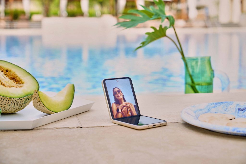 Photo d’une Samsung Galaxy Z Flip3 repliee a la moitie sur une table pres d’une assiette avec un melon, au bord de l’eau 