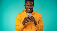 Photo sur fond turquoise d’un jeune homme noir souriant a la camera tout en tenant sons smartphone entre les deux mains