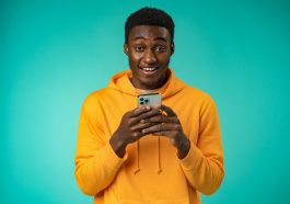 Photo sur fond turquoise d’un jeune homme noir souriant a la camera tout en tenant sons smartphone entre les deux mains