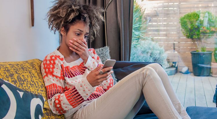 Une femme assise sur le sofa l’air choque en regardant son smartphone