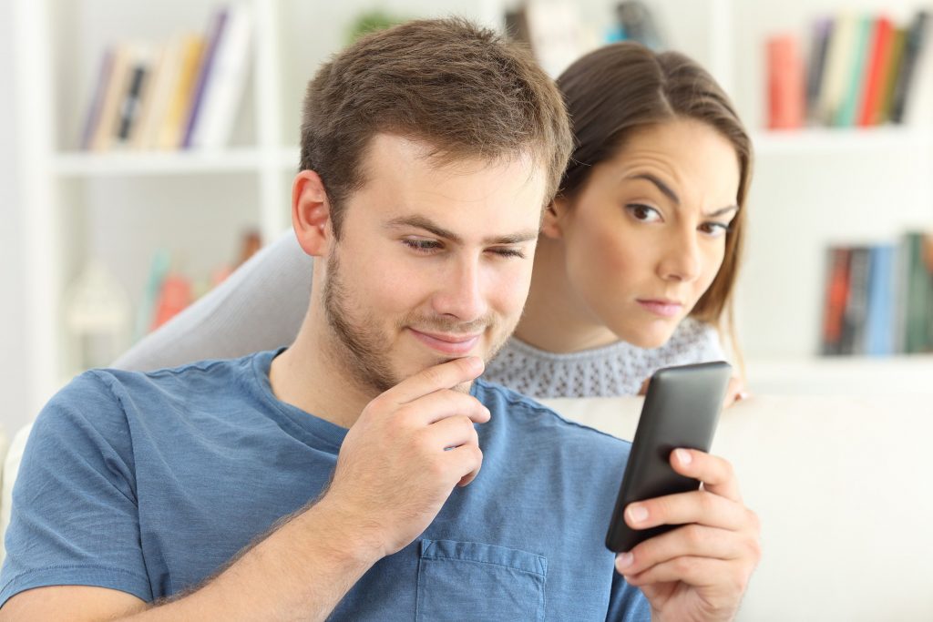 Un homme regardant son smartphone et une femme se tenant derriere lui espionnant ce qu’il fait