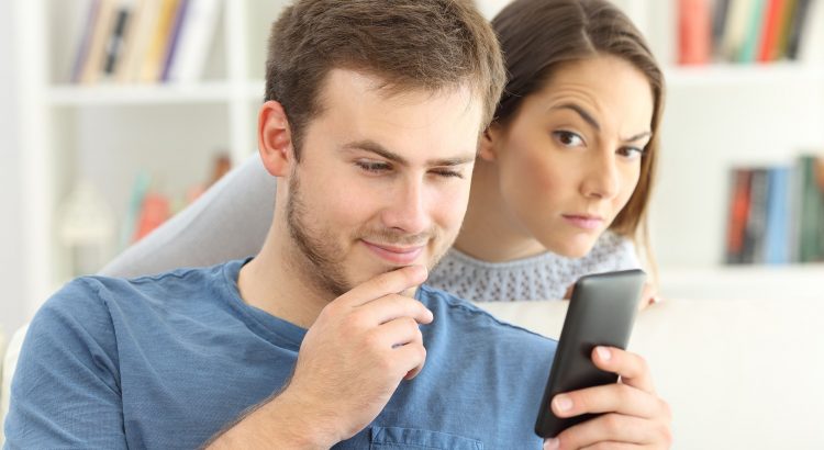 Un homme regardant son smartphone et une femme se tenant derriere lui espionnant ce qu’il fait
