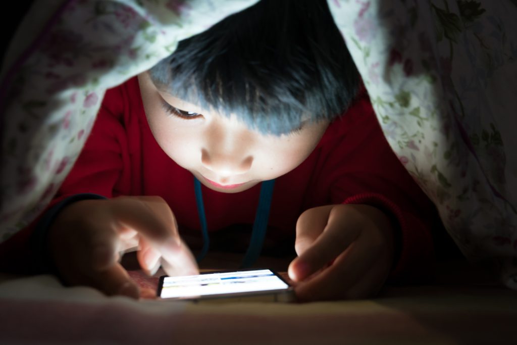 Un petit garcon asiatique jouant a un smartphone sous la couverture