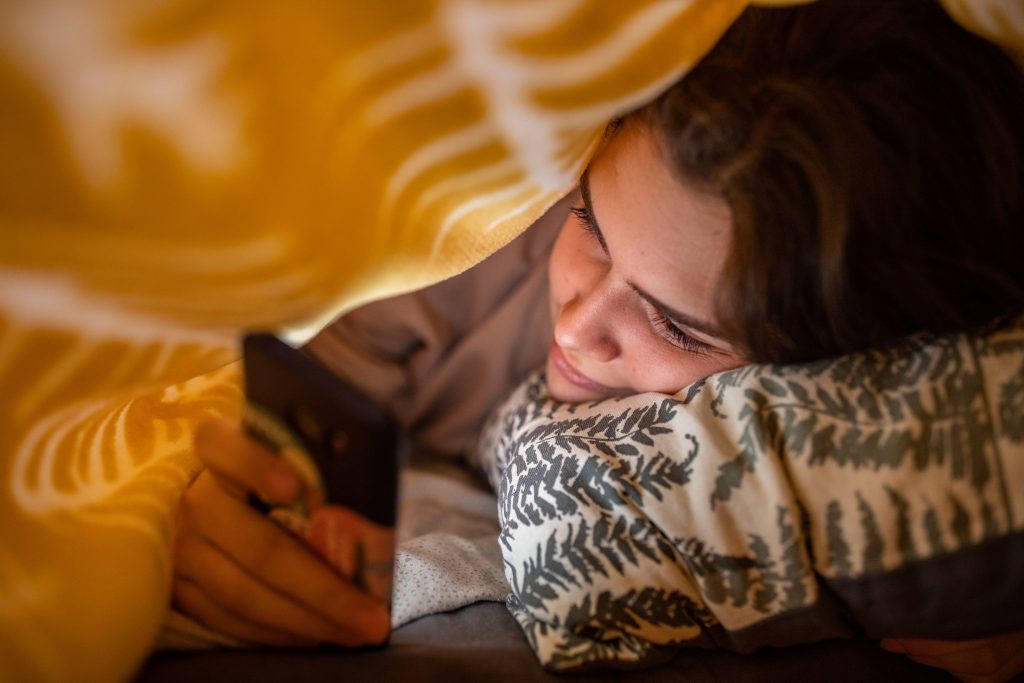Une jeune femme scrollant son smartphone sous une couverture